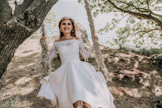 Young european beautiful girl in white bridal marriage dress\
posing on swing in forest trees on the ocean sea beach background\
exotic wedding photography ideas