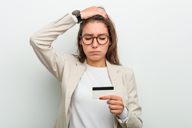 Young european business woman holding a credit card being shocked, she has remembered important meeting.