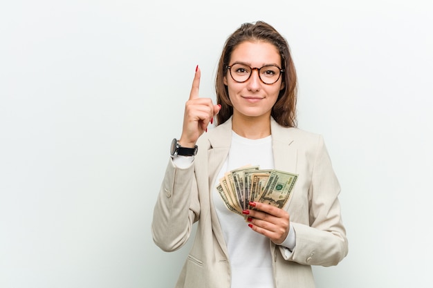 Young european business woman holding dollar banknotes showing number one with finger