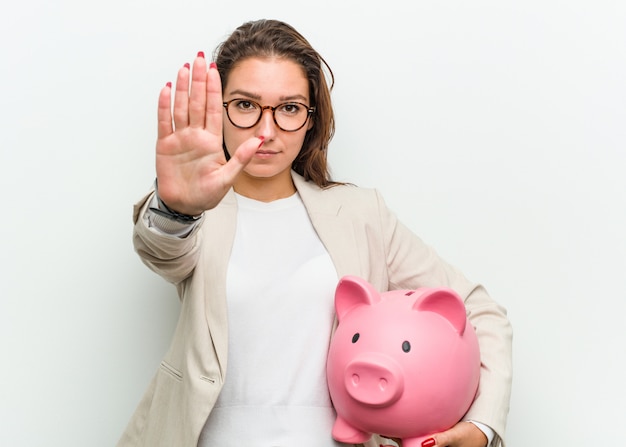 Young european business woman holding a piggy bank standing with outstretched hand showing stop sign, preventing you.