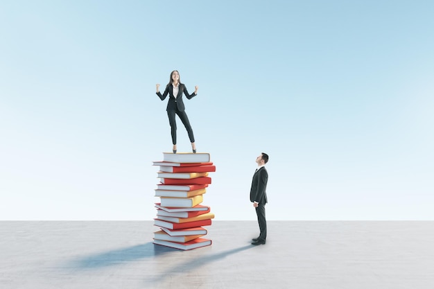 Young european businessman looking up at happy female colleague\
standing on book pile blue sky background education knowledge and\
success concept