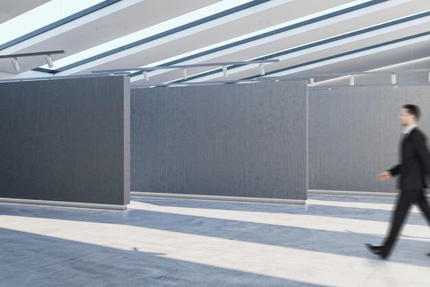 Giovane uomo d'affari europeo che cammina nell'interno moderno della sala espositiva in cemento con un posto vuoto sulla parete e la luce del sole