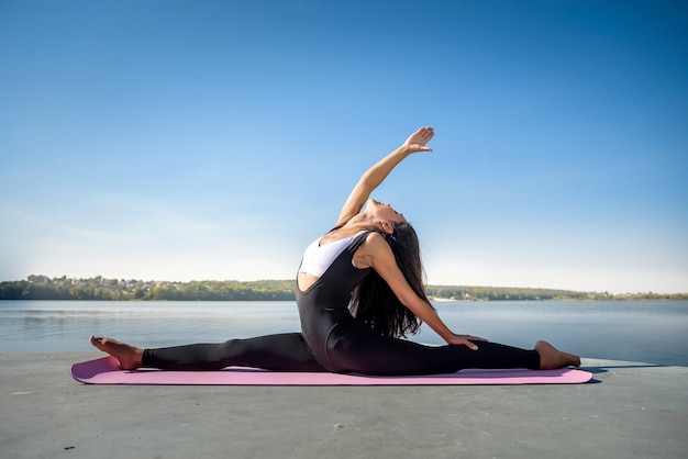 Young european female athlete  doing exercises for stretching the legs, sits on the splits outside, near the lake. Healthy lifestyle