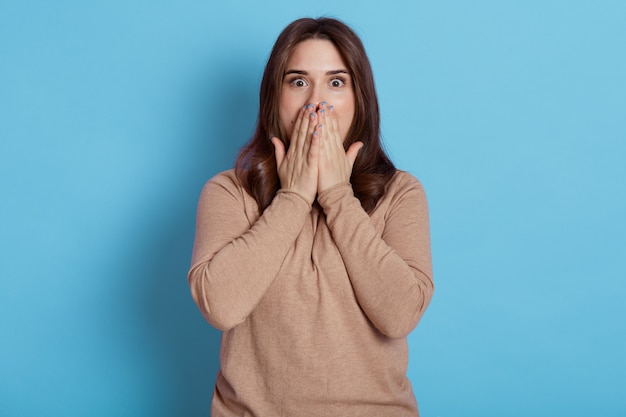 Young European female dresses beige shirt, looking with fear in eyes, covering mouth with palms, looks with big eyes, posing isolated over blue wall.