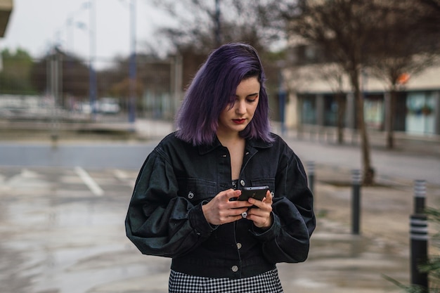 Foto giovane donna europea con i capelli viola che cammina per strada e scrive messaggi sul suo telefono