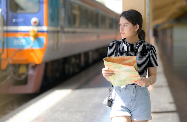 A young European girl traveler holding a map standing on the train platform, with a camera and headphones, leaning on a pole on the platform and looking at the train, train journey.