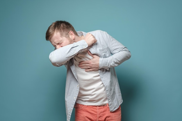 Young European man coughs at elbow and holds hand on chest experiencing pain on a blue background