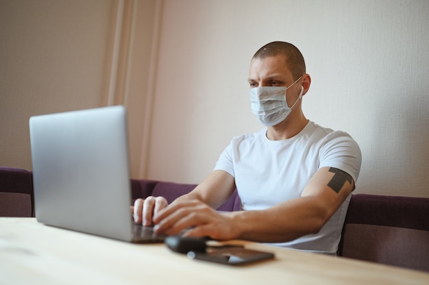 Young european man in face protective medicine mask working on a laptop during coronavirus isolation