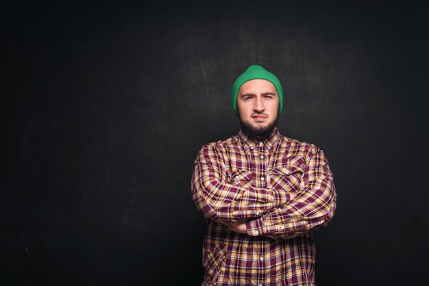 Молодой европейский мужчина с бородой в зеленой вязаной шапке выглядит удивленным и озадаченным. Показывает пальцами вверх и в правую сторону. Черный фон, пустая копия пространства для текста или рекламы