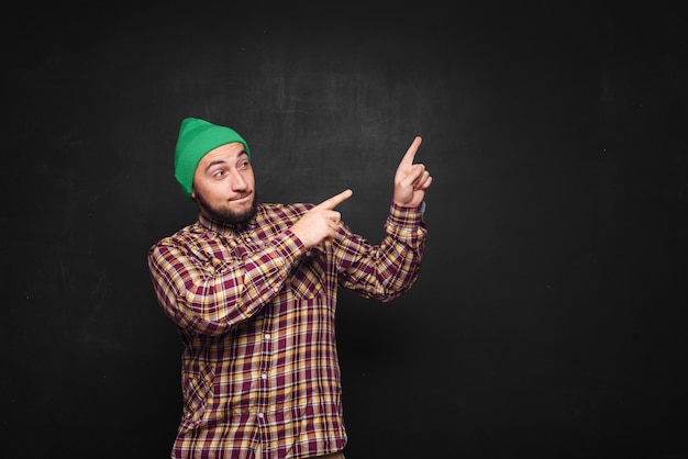 Photo young european man with beard in green knitted hat , looks surprised and puzzled. showing fingers upwards and right side. black background, blank copy space for text or advertisement