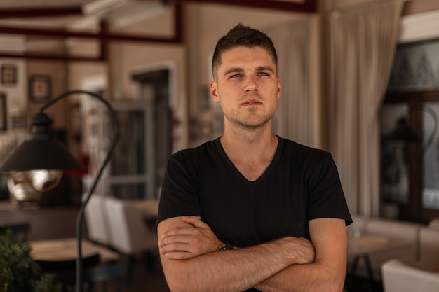 Young european man with stylish hairstyle in a black t-shirt stands in a vintage restaurant