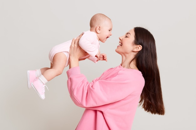 Giovane madre europea che tiene la sua bambina in mano e guarda il suo bambino con amore e sorriso gentile, femmina con i capelli scuri che indossa un maglione casual rosa, giocando con il suo bambino.