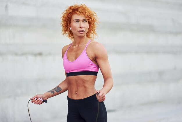 Young european redhead woman in sportive clothes jumping with rope outdoors