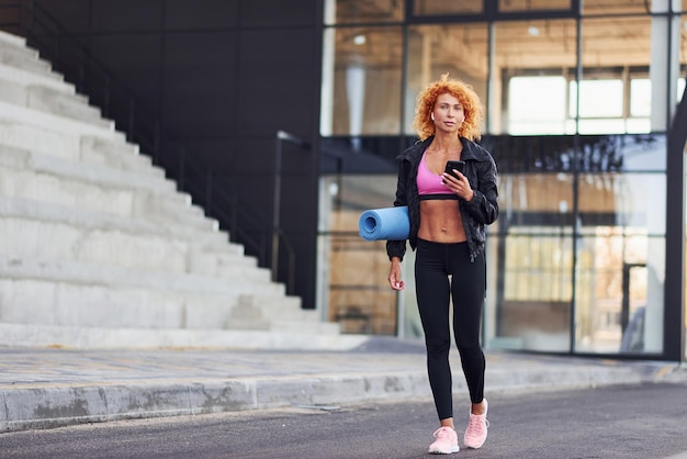 Foto giovane donna rossa europea in abiti sportivi con telefono e tappetino in mano all'aperto