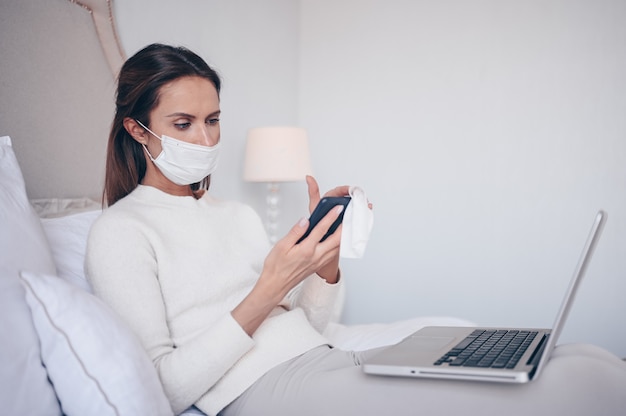 Young european woman in face mask in bedroom with laptop during coronavirus isolation