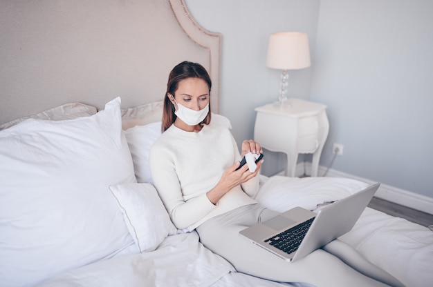 Young european woman in face mask in bedroom with laptop during coronavirus isolation