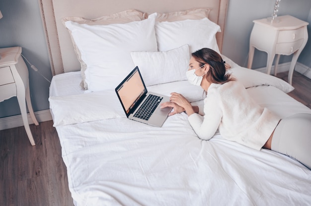 Young european woman in face medicine mask working on a laptop in bedroom during coronavirus isolation