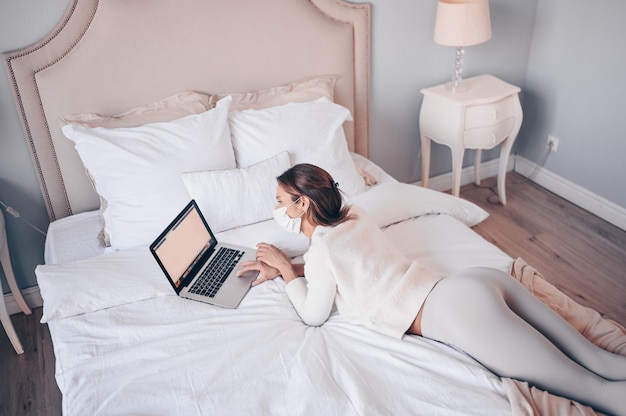 Young european woman in face medicine mask working on a laptop in bedroom during coronavirus isolation