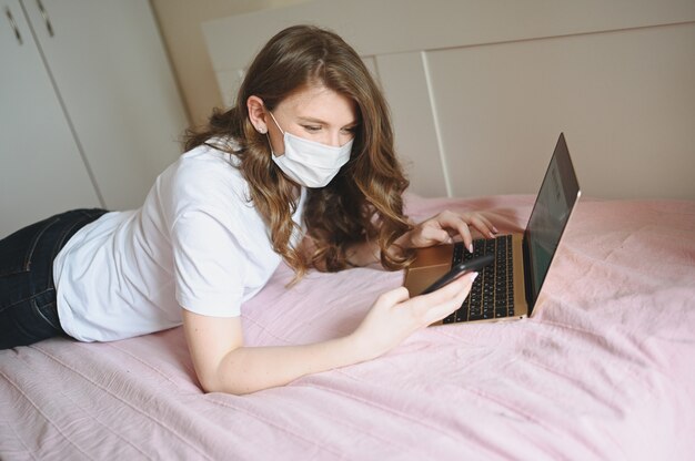 Young european woman in face medicine mask working on a laptop and phone in bed