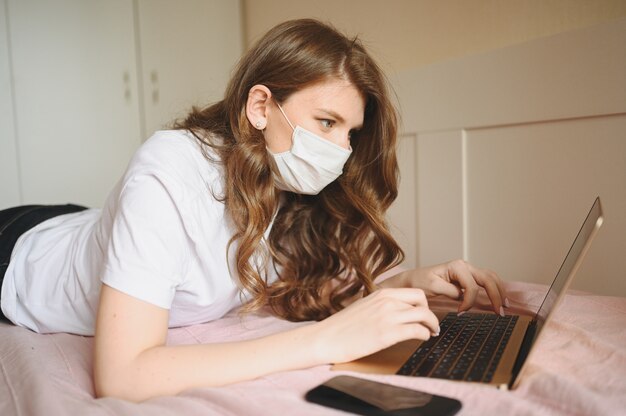 Young european woman in face medicine mask working on a laptop and phone in bed