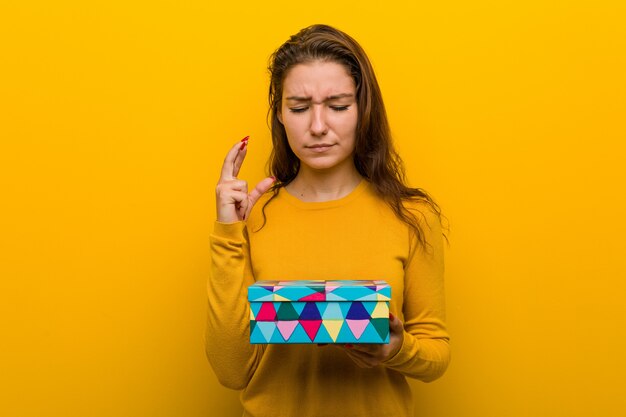 Young european woman holding a gift crossing fingers for having luck