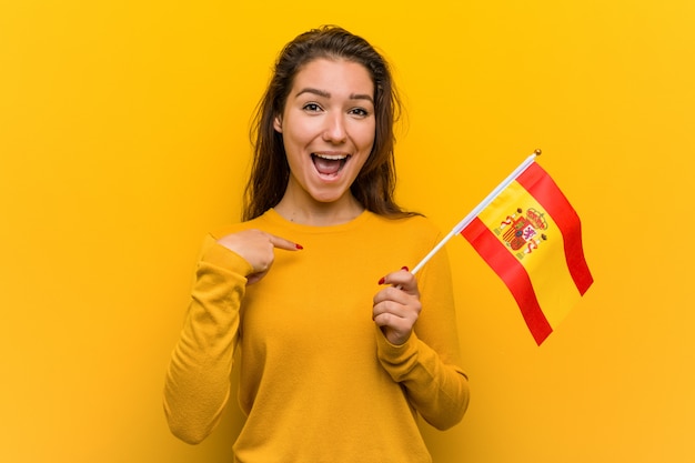 Foto la giovane donna europea che tiene una bandiera spagnola ha sorpreso indicare se stessa, sorridendo largamente.