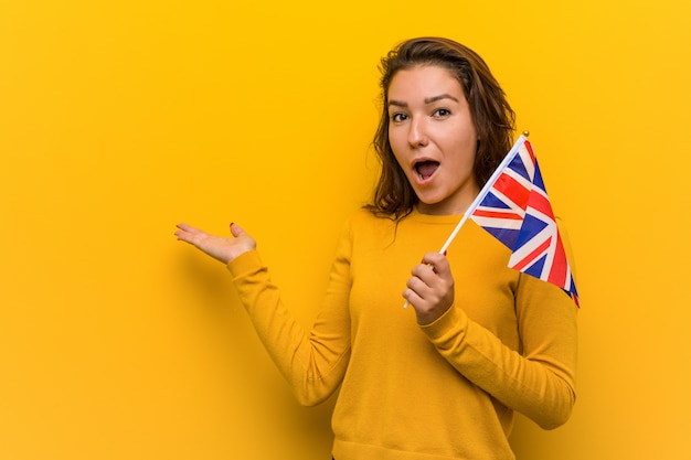 Young european woman holding an united kingdom flag impressed holding copy space on palm