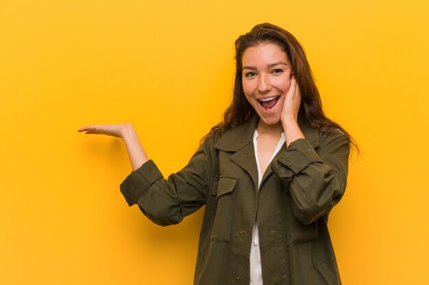 Young european woman isolated over yellow holds copy space on a palm, keep hand over cheek. Amazed and delighted.