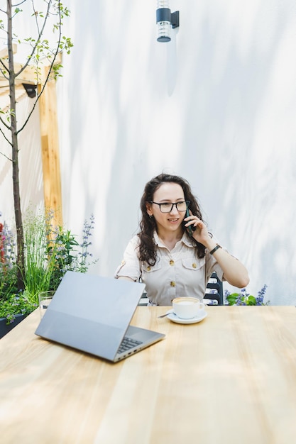 ノートパソコンとコーヒーを飲みながら屋外のカフェテーブルに座るヨーロッパの若い女性は、カフェで在宅勤務を楽しんだり、オンラインで勉強したりする眼鏡をかけた笑顔の女性