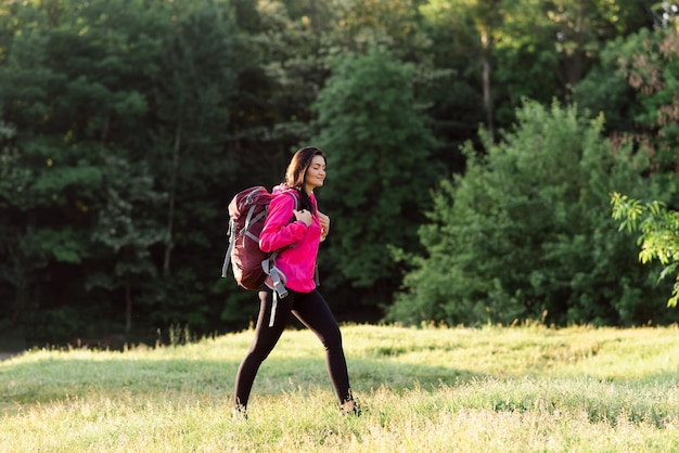 Young european woman walking with backpack on green meadow near forest Beautiful smiling girl wear sportswear and pink jacket Concept of resting and tourism on nature Sunny daytime