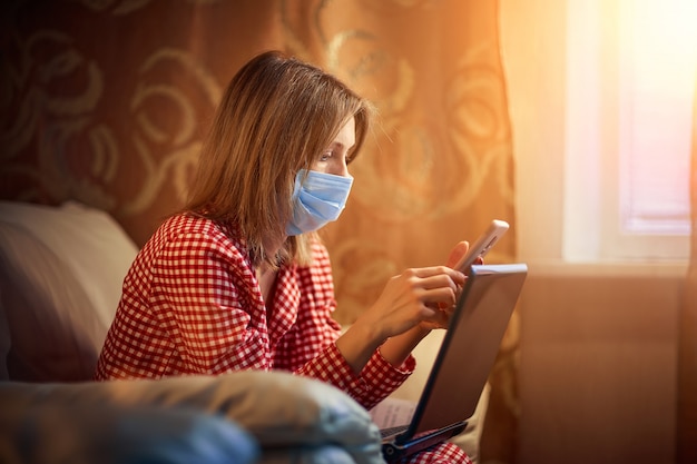 Foto la giovane donna europea che indossa la maschera chirurgica resta a casa durante il coronavirus, l'epidemia covid-19 e guarda notizie o applicazioni sullo smartphone.