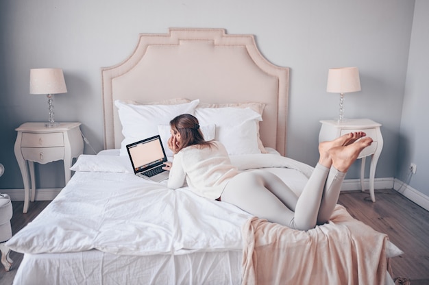Молодая европейская женщина работает на ноутбуке в белой спальне
