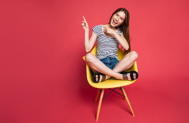 Young excited beautiful blonde woman in casual wear is sitting on yellow chair in big studio with pink background. Happy and funny stylish girl