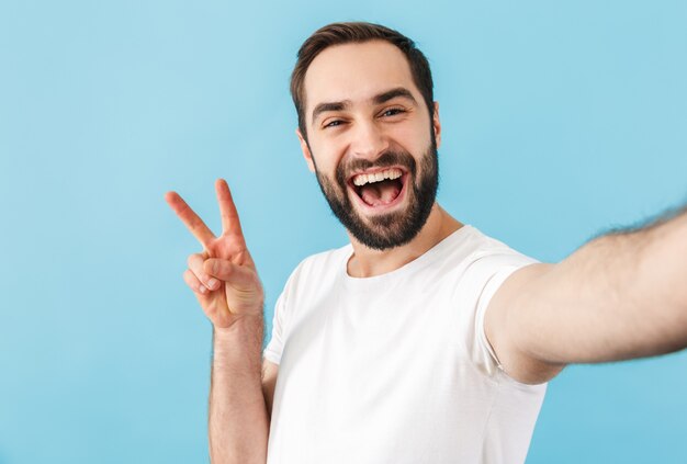 young excited cheerful man isolated over blue wall take a selfie by camera showing peace gesture.