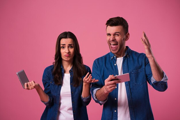 Young excited couple standing isolated over pink background in casual wear, playing games on mobile phones