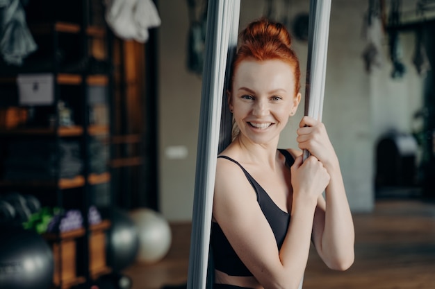 Young excited ginger female with hair in bun posing in fitness studio, sitting in grey aerial yoga hammock and looking into camera with smile, wearing black sportswear. Anti gravity yoga class concept