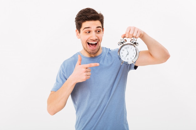 Young excited man in casual t-shirt and holding and pointing finger on alarm clock, isolated over white wall