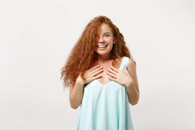 Giovane ragazza sorridente emozionante della donna della testarossa in vestiti leggeri casuali che posano isolato su fondo bianco in studio concetto di stile di vita di emozioni sincere della gente. mock up copia spazio. tenendo le mani sul petto.