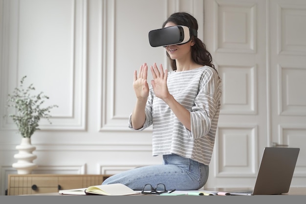 Young excited woman freelancer sitting on top of desk at home office wearing vr glasses