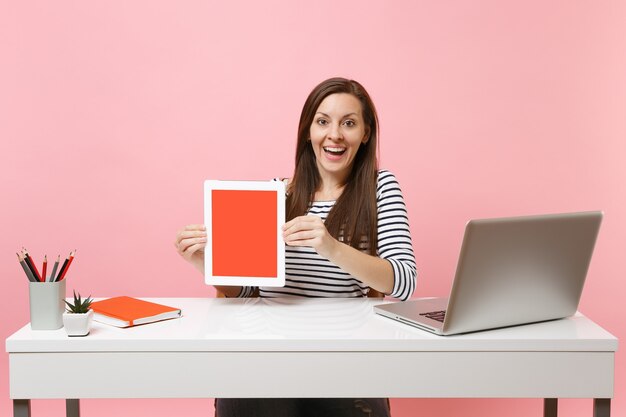 Young excited woman hold tablet computer with blank empty screen sit work at white desk with contemporary pc laptop isolated on pastel pink background. Achievement business career concept. Copy space.