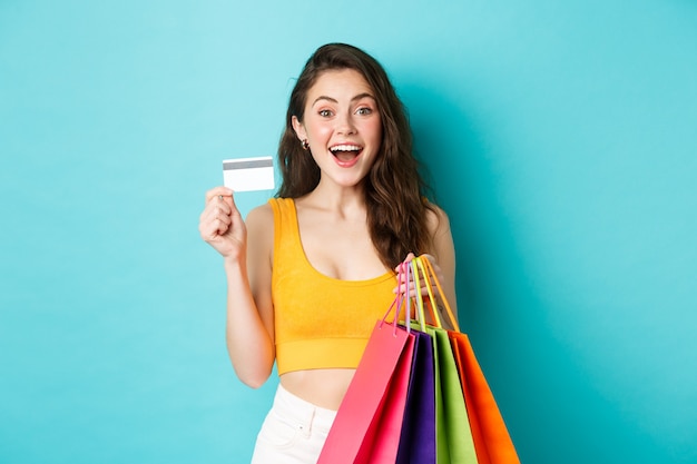 Giovane donna eccitata che tiene le borse della spesa e mostra la carta di credito in plastica, compra cose durante le offerte promozionali, in piedi su sfondo blu.
