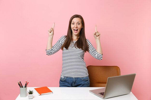 Young excited woman pointing index fingers up work and standing near white desk with pc laptop 