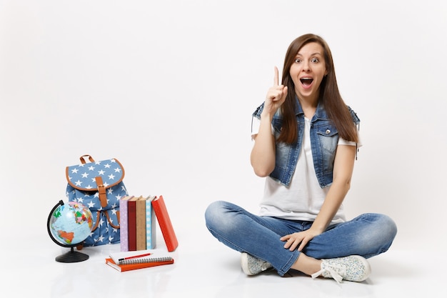 Young excited woman student enlightened with new thought, idea pointing index finger up sit near globe, backpack school books isolated on white wall