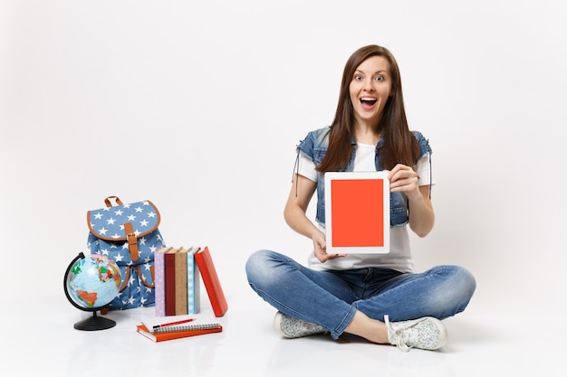 Young excited woman student holding tablet pc computer with blank black empty screen sitting near globe backpack school books isolated