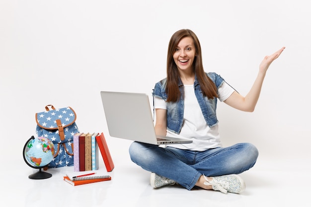 Young excited woman student holding using laptop pc computer spreading hand sitting near globe backpack, school books isolated on white wall