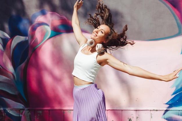 Photo a young excited woman in a top and skirt wearing headphones listening to music walking dancing with her hands up in the open air against a pink wall