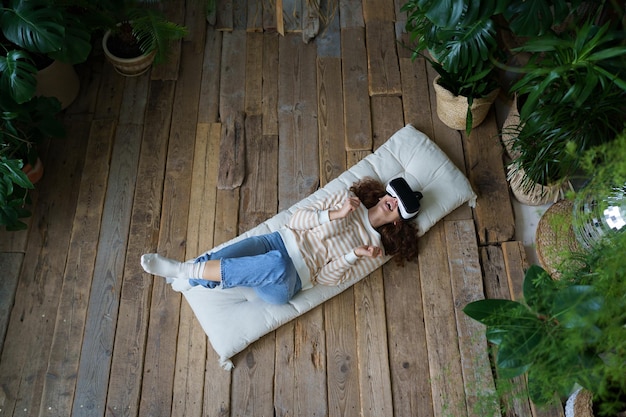 가상 현실을 즐기며 매트리스에 누워 온실에서 쉬고 있는 VR 헬멧을 쓴 젊은 흥분한 여성