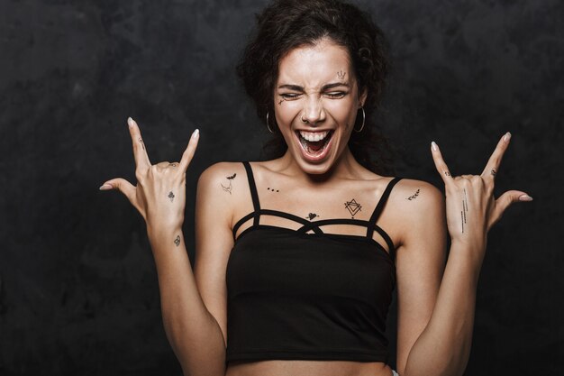 Giovane donna eccitata con tatuaggio che urla e gesticola le corna con le dita isolate su un muro nero