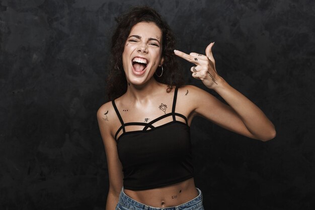 young excited woman with tattoo smiling and showing middle finger isolated over black wall