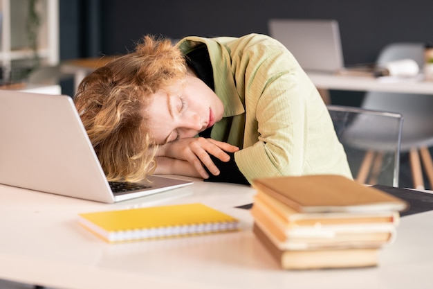 Young exhausted female student or office manager keeping head on hands while napping in front of laptop with stack of books near by